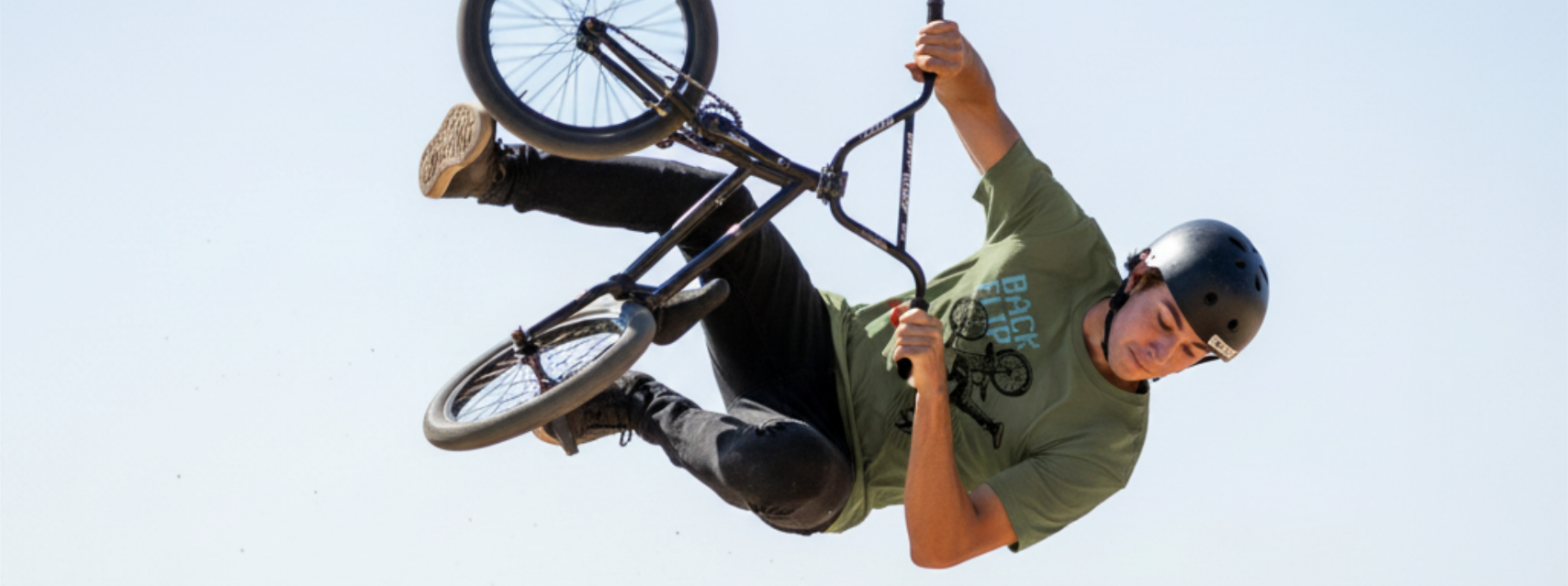 Ein BMX-Fahrer führt in der Luft über einer Dirt-Jump-Schanze einen Trick aus. Er trägt ein olivgrünes T-Shirt mit der Aufschrift „BACK FLIP“, eine schwarze Hose, einen schwarzen Helm und braune Sneaker. Das Fahrrad ist fast senkrecht aufgerichtet, während der Fahrer seitlich versetzt zum Rahmen in der Luft hängt. Der Hintergrund zeigt einen klaren, hellen Himmel und unscharfe Bäume am Horizont.